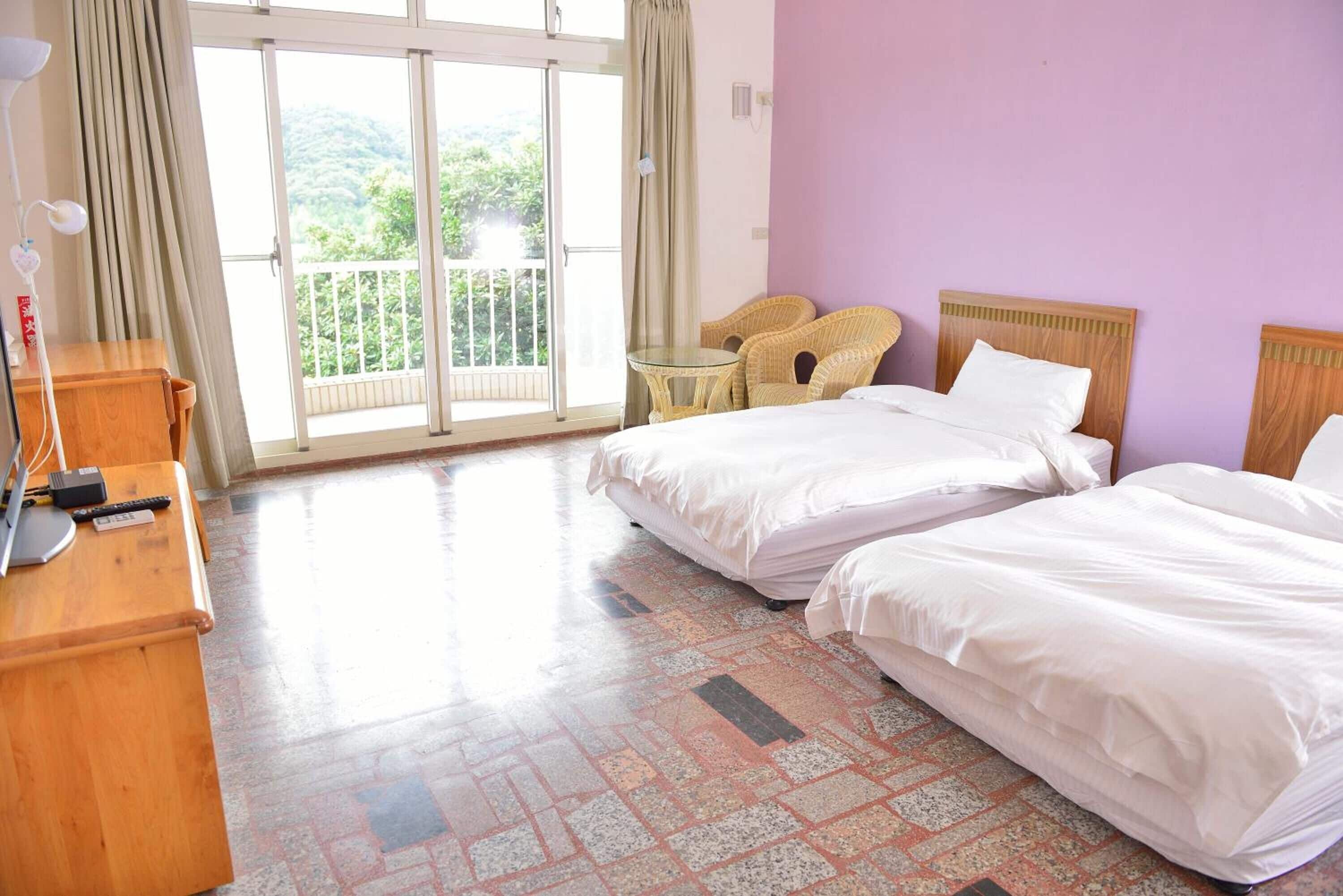 A hotel room with two beds, a wooden desk, a small round table, and a balcony with a view of greenery.