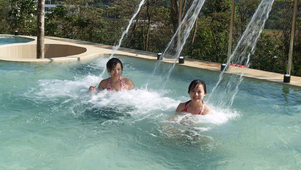 A swimming pool with water jets, surrounded by greenery and buildings in the background. - Wan Jin Hot Spring (New Taipei City)