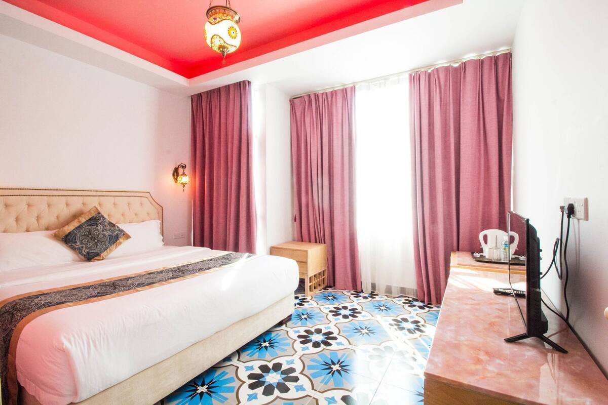 A hotel room with a bed, a TV, a small table, and a window with curtains.