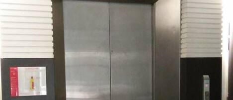An elevator with a metallic finish, a control panel on the right, and a fire alarm on the left.