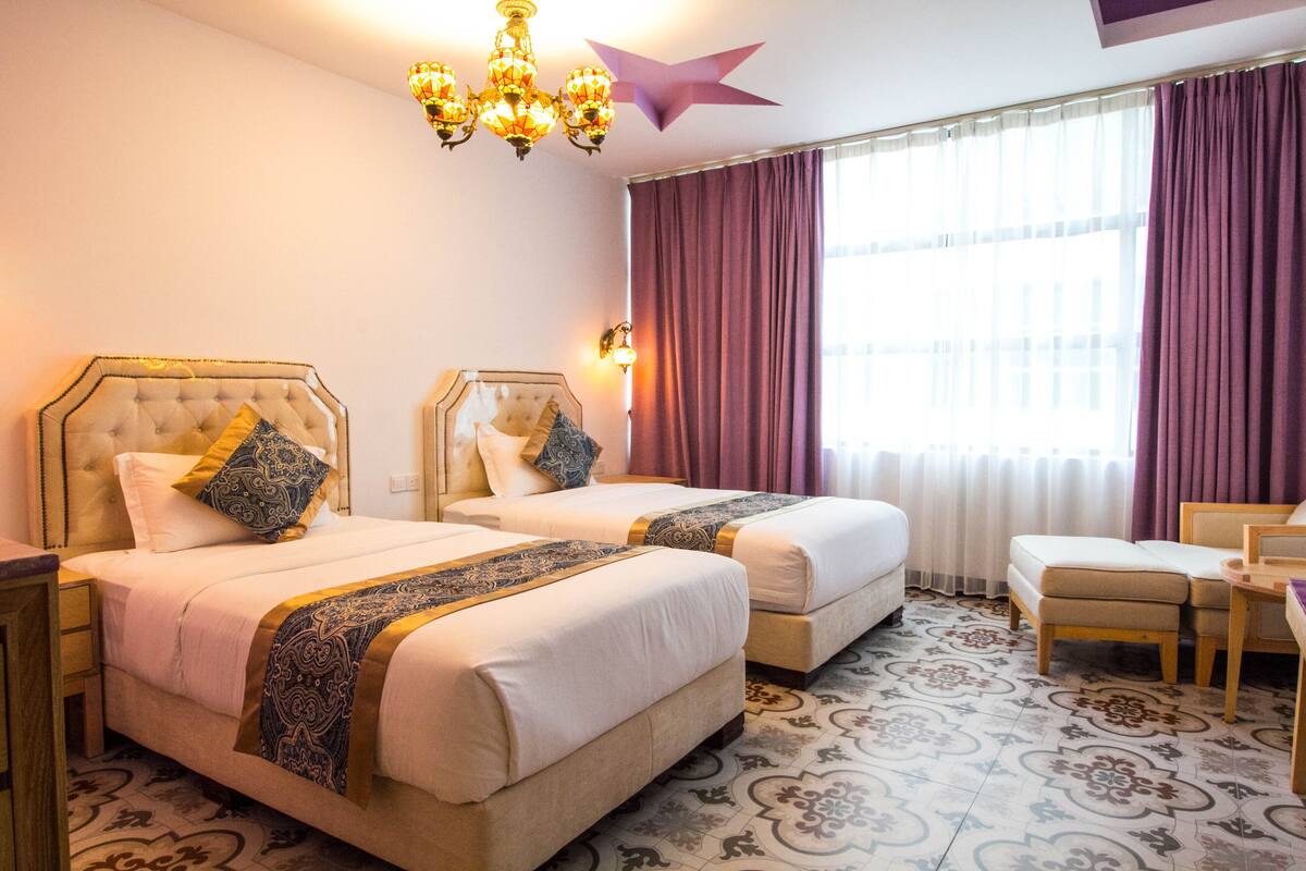 A hotel room with two beds, a chandelier, a star decoration, a window with curtains, and a patterned floor.