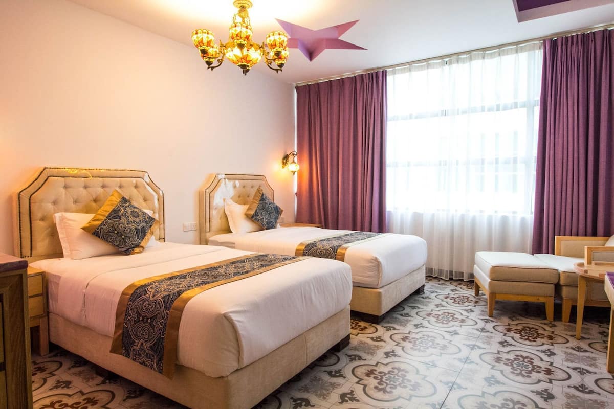 A hotel room with two beds, a chandelier, a star decoration, a window with curtains, and a small desk with a chair.