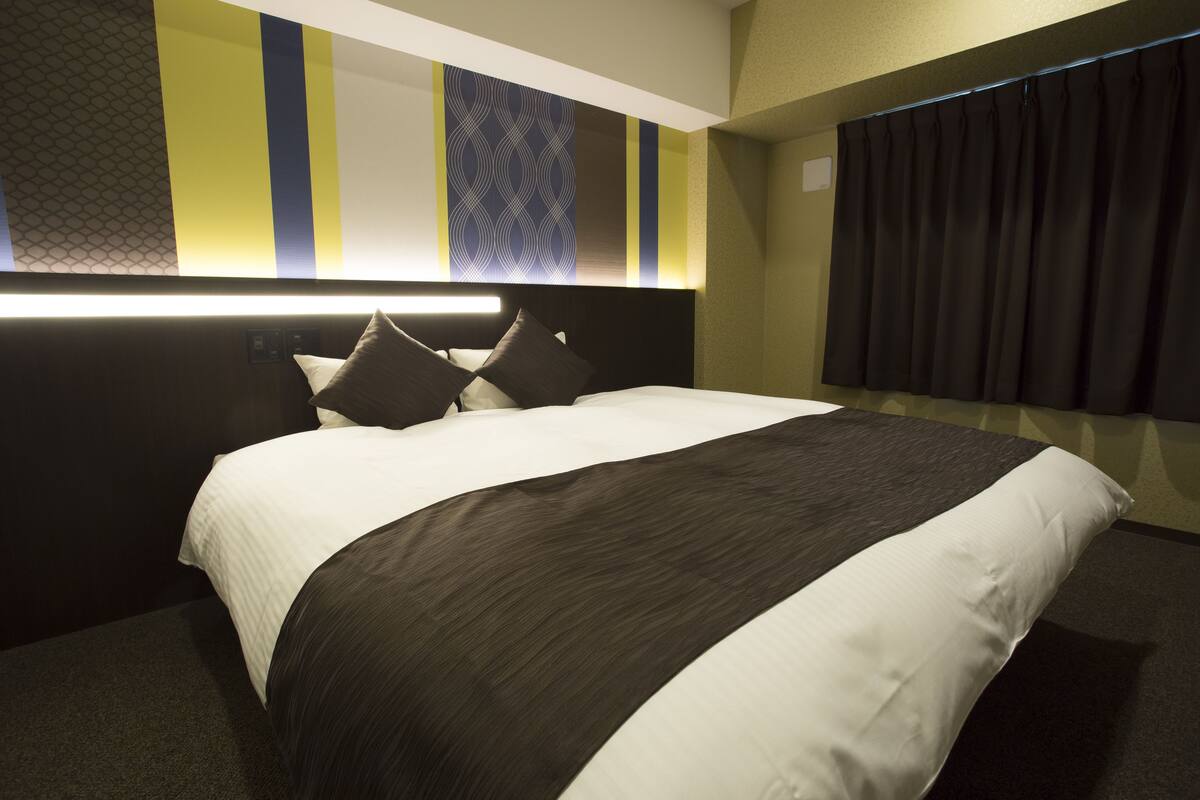 Une chambre d’hôtel avec un grand lit, une tête de lit dotée d’un éclairage intégré, un mur orné d’un motif et un rideau foncé à la fenêtre.
