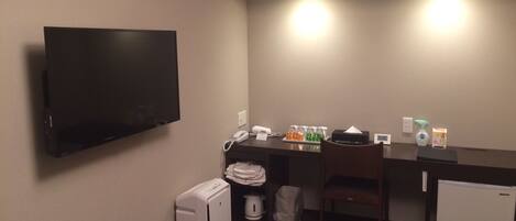A hotel room with a flat-screen TV, a desk with a phone, a small fridge, a trash bin, and a heater.