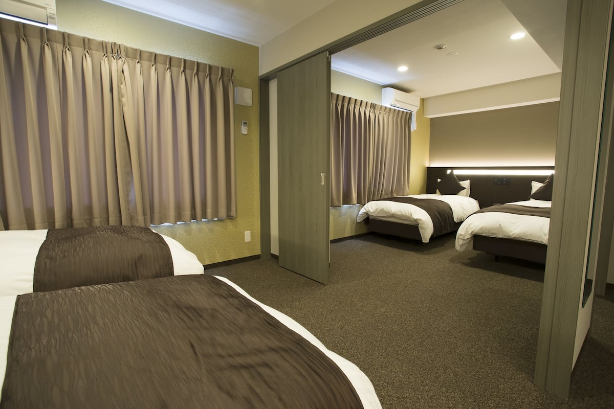 Une chambre d’hôtel avec deux lits, une grande fenêtre avec des rideaux, une table de chevet et une tête de lit dotée d’un éclairage intégré.