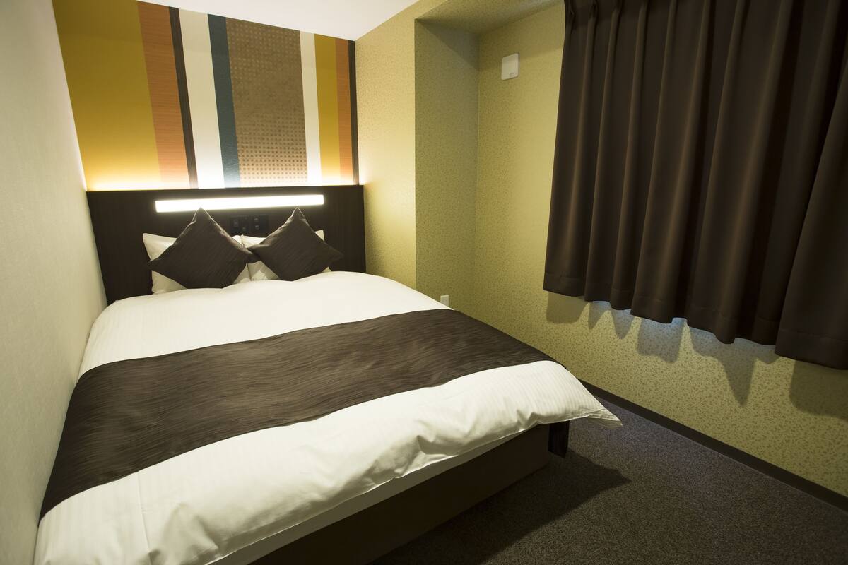 Une chambre d’hôtel avec un lit, une tête de lit, une lampe fixée au mur et une fenêtre avec des rideaux.