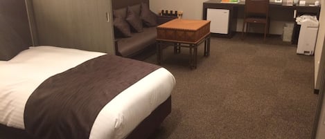A hotel room with a large bed, a sofa, a small table, a desk, and a refrigerator.