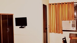 A room with a wooden wardrobe, a flat-screen TV mounted on the wall, a window with golden curtains, and an air conditioning unit. - Accent Hotel (Greater Noida)