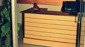 A reception area with a sign labeled "RECEPTION," a counter with drawers, a small statue, and a plant to the left. - Accent Hotel (Greater Noida)