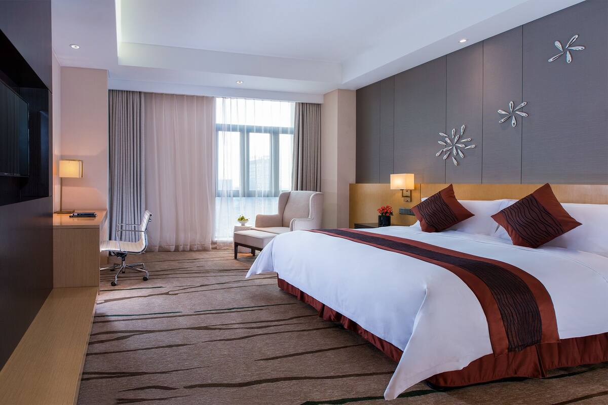 Ein Hotelzimmer mit einem großen Bett, einem Schreibtisch mit Stuhl, einem Fernseher und einem Fenster mit Vorhängen.