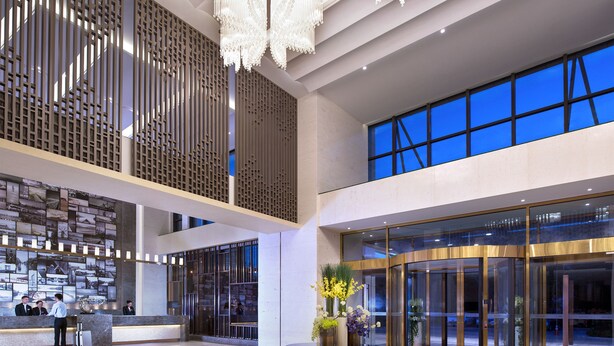 Um hall amplo com teto alto, lustres modernos, um balcão de recepção e uma grande janela com vista para o céu noturno.
