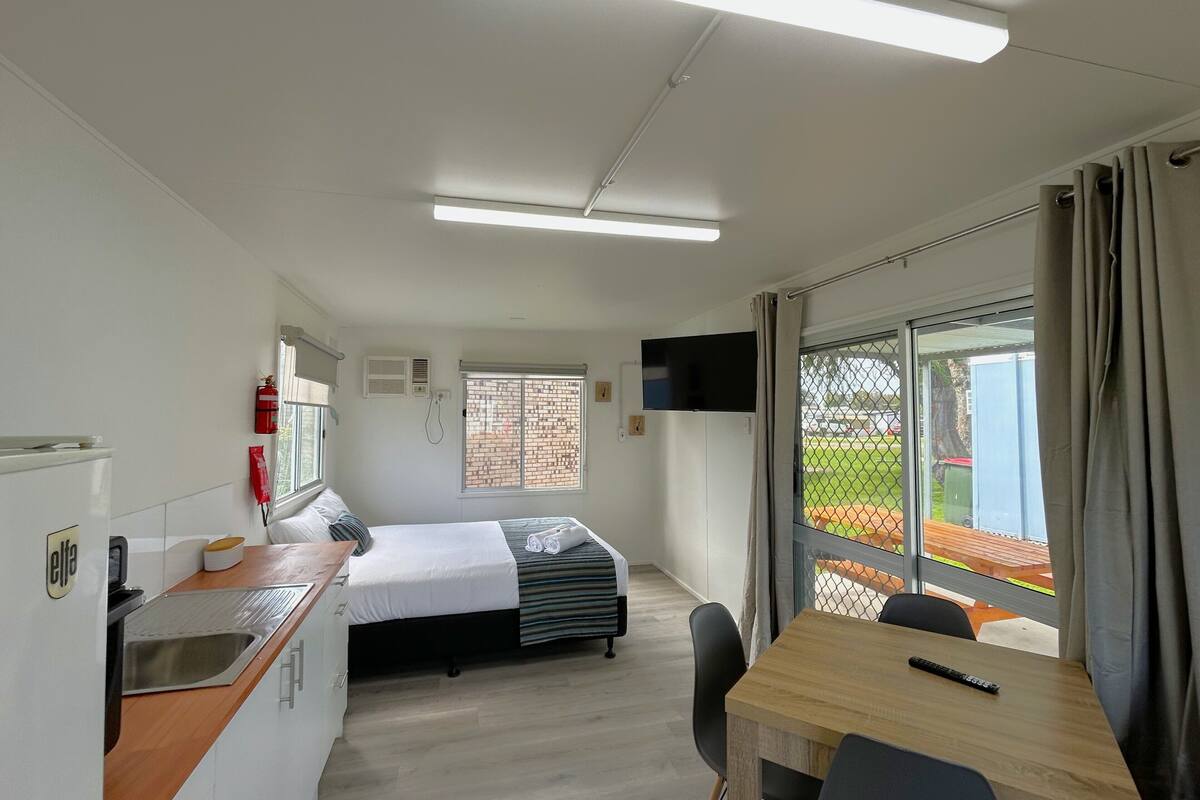 A compact room with a bed, a small kitchenette, a dining area, and a large window with a view of the outdoors.
