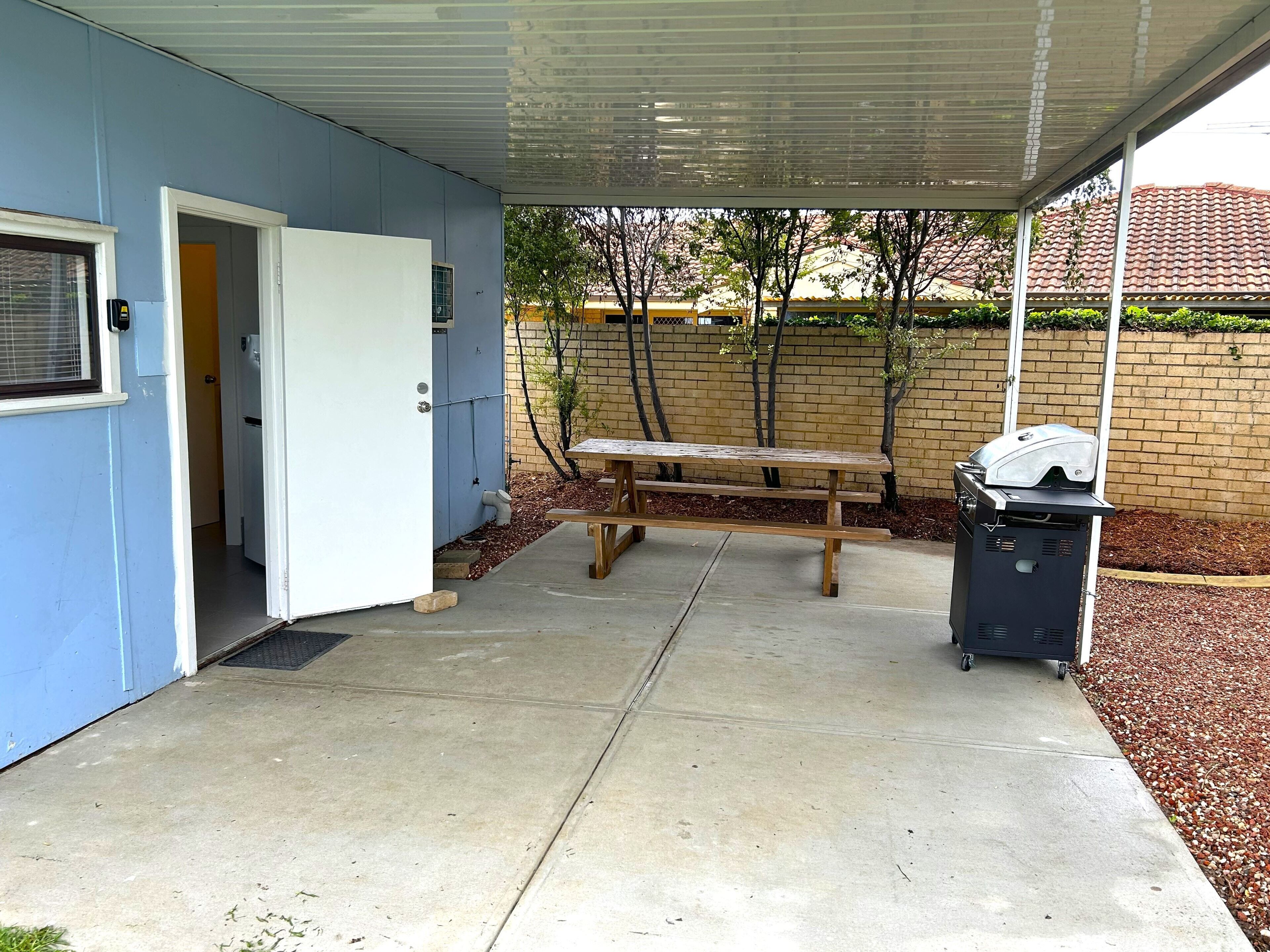 A covered patio area with a concrete floor, a wooden bench, a barbecue grill, and a door leading to an interior space.