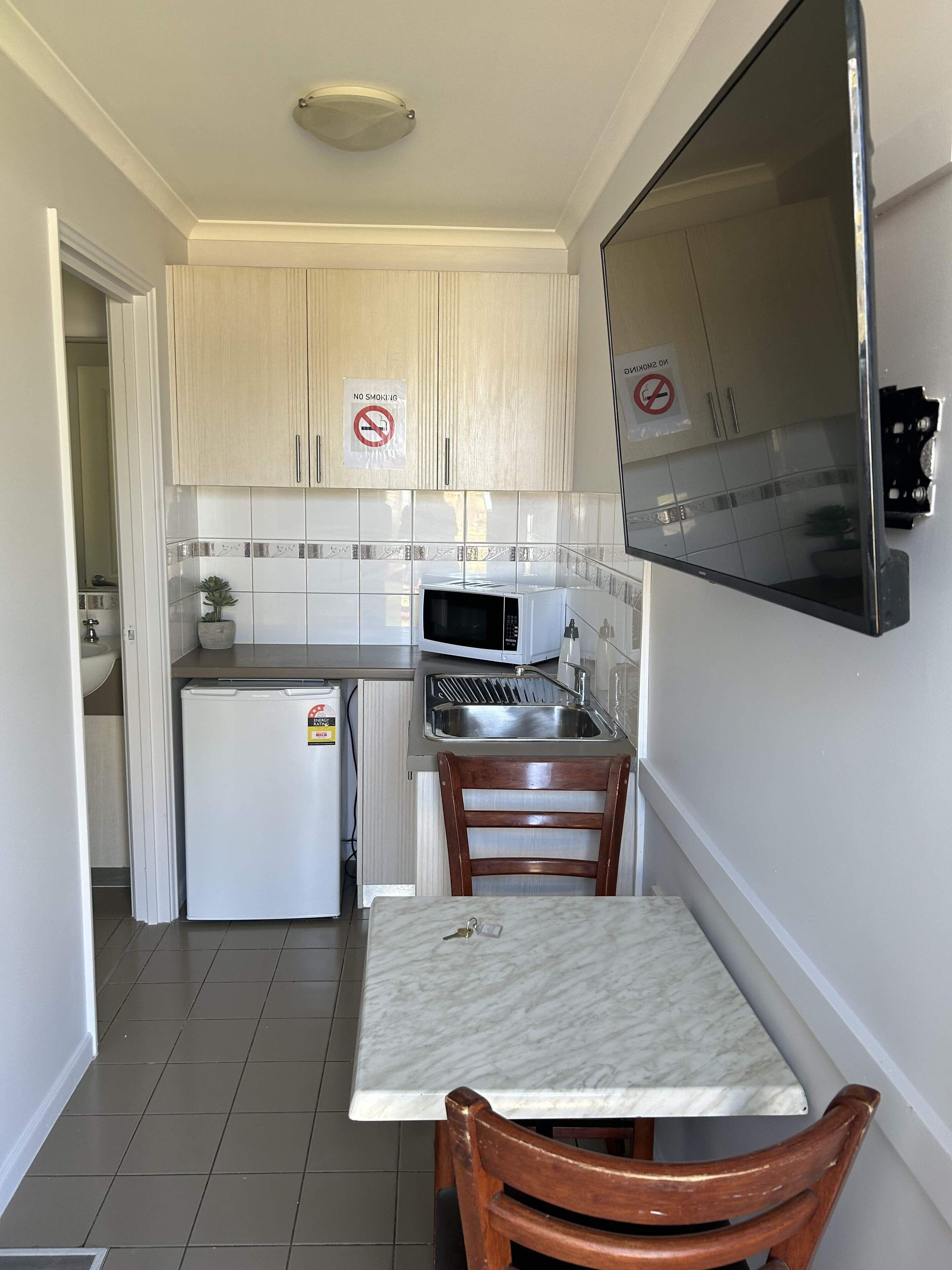 A compact kitchen with white cabinets, a microwave, a sink, and a small refrigerator. There is a wooden chair and a table with a key on it.