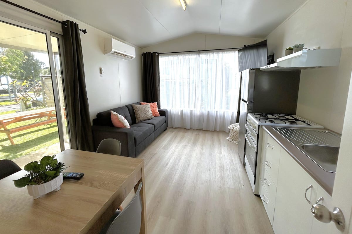 A compact living space with a kitchenette, a sofa with cushions, a dining table with a plant, and a view of the outdoors through a large window.