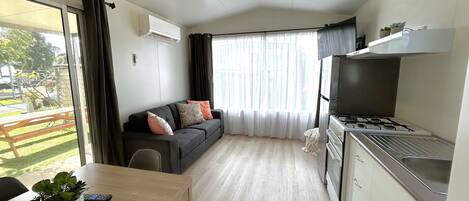 A compact living space with a kitchenette, a sofa with cushions, a dining table with a plant, and a view of the outdoors through a large window.