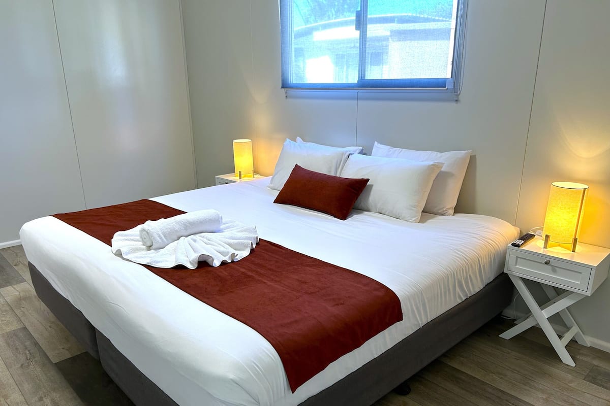 A neatly made bed with white and red linens, a remote control, and a bedside lamp on a white nightstand.