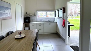 A compact kitchen with white cabinets, a wooden dining table, and a window overlooking a grassy area. - Mandurah Coastal Holiday Park (Mandurah)
