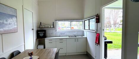 Cocina compacta con muebles blancos, una mesa de comedor de madera y ventana con vista a un área con césped.