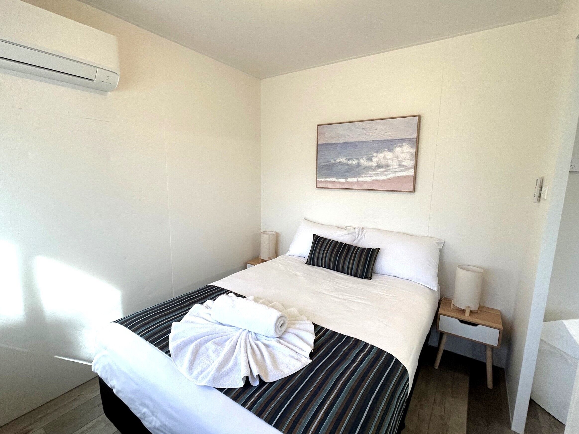 A neatly made bed with a striped bedspread, a rolled towel, and a pillow. There is a painting of a beach scene on the wall, a bedside table with a lamp, and an air conditioning unit mounted on the wall.