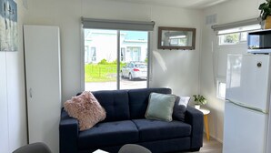 A modern living room with a dark blue sofa, a fluffy pillow, and a small table. There is a white refrigerator and a microwave on top of it. A window with a view of a car and a building is visible. - Mandurah Coastal Holiday Park (Mandurah)
