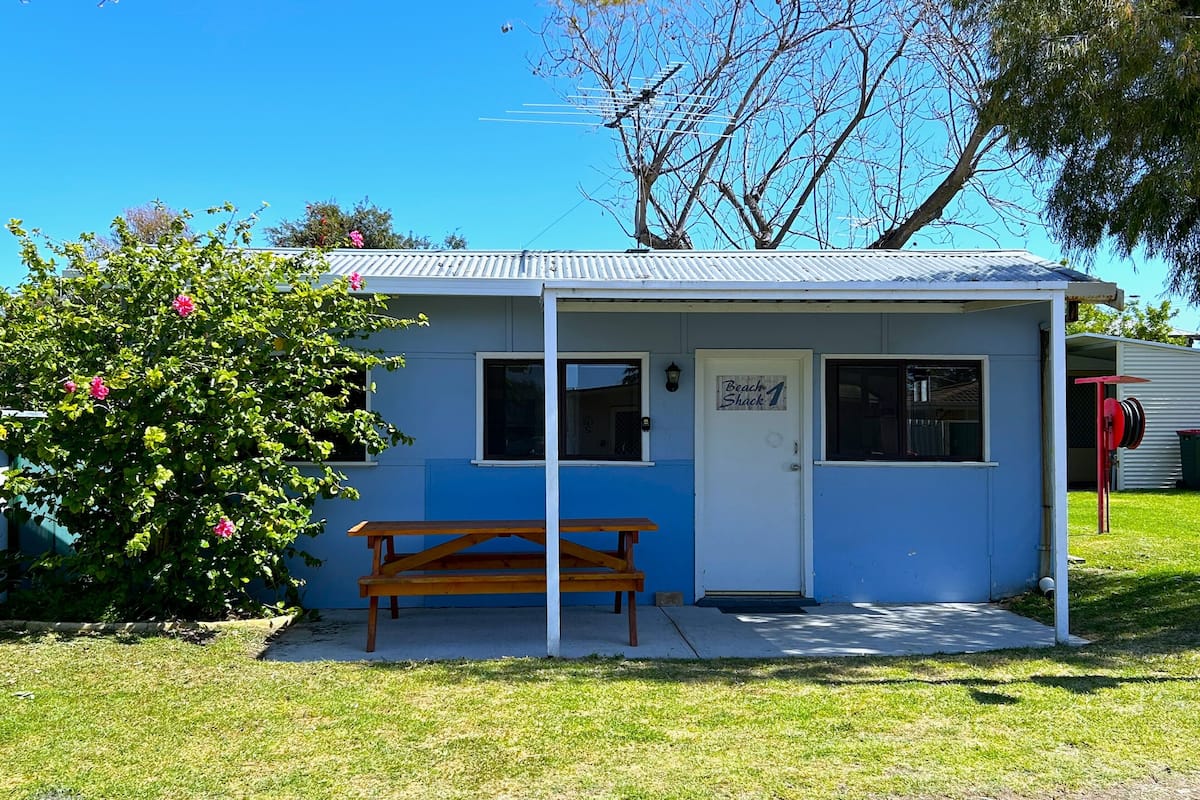 A small, single-story building with a blue exterior and a white door. There is a wooden bench outside and a sign that reads "Beach Shack."