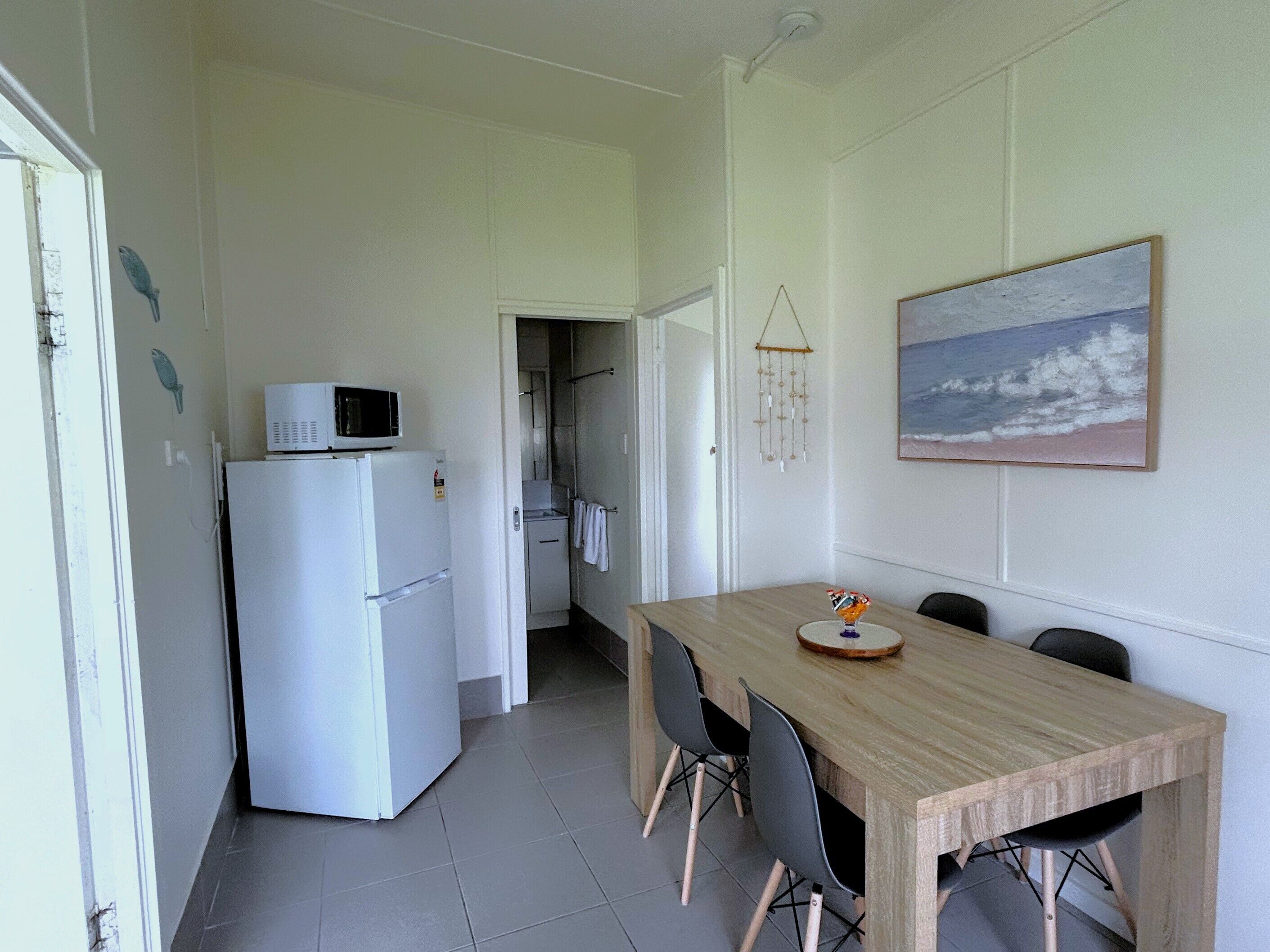 Een compacte keuken met een witte koelkast, een magnetron en een eethoek met een houten tafel en vier stoelen. Op de muur hangt een schilderij van een strand.