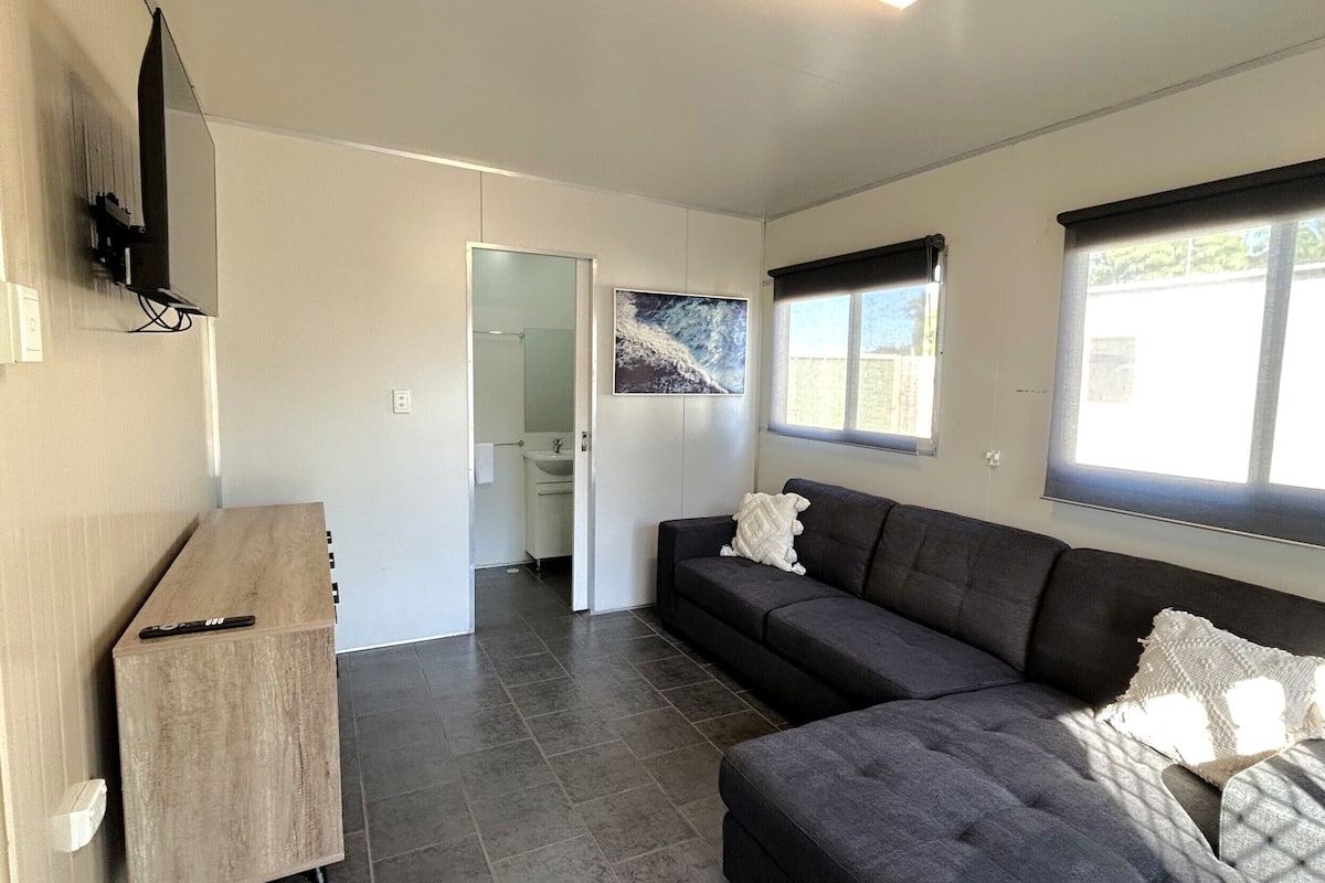 A modern living room with a grey sofa, a wooden cabinet, a flat-screen TV mounted on the wall, and a window with blinds.