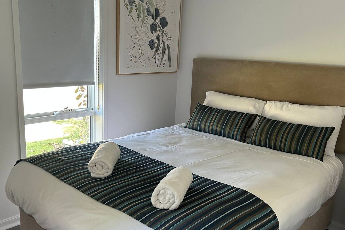 A neatly made bed with striped bedding and two rolled towels, a headboard, and a window with a view of greenery.