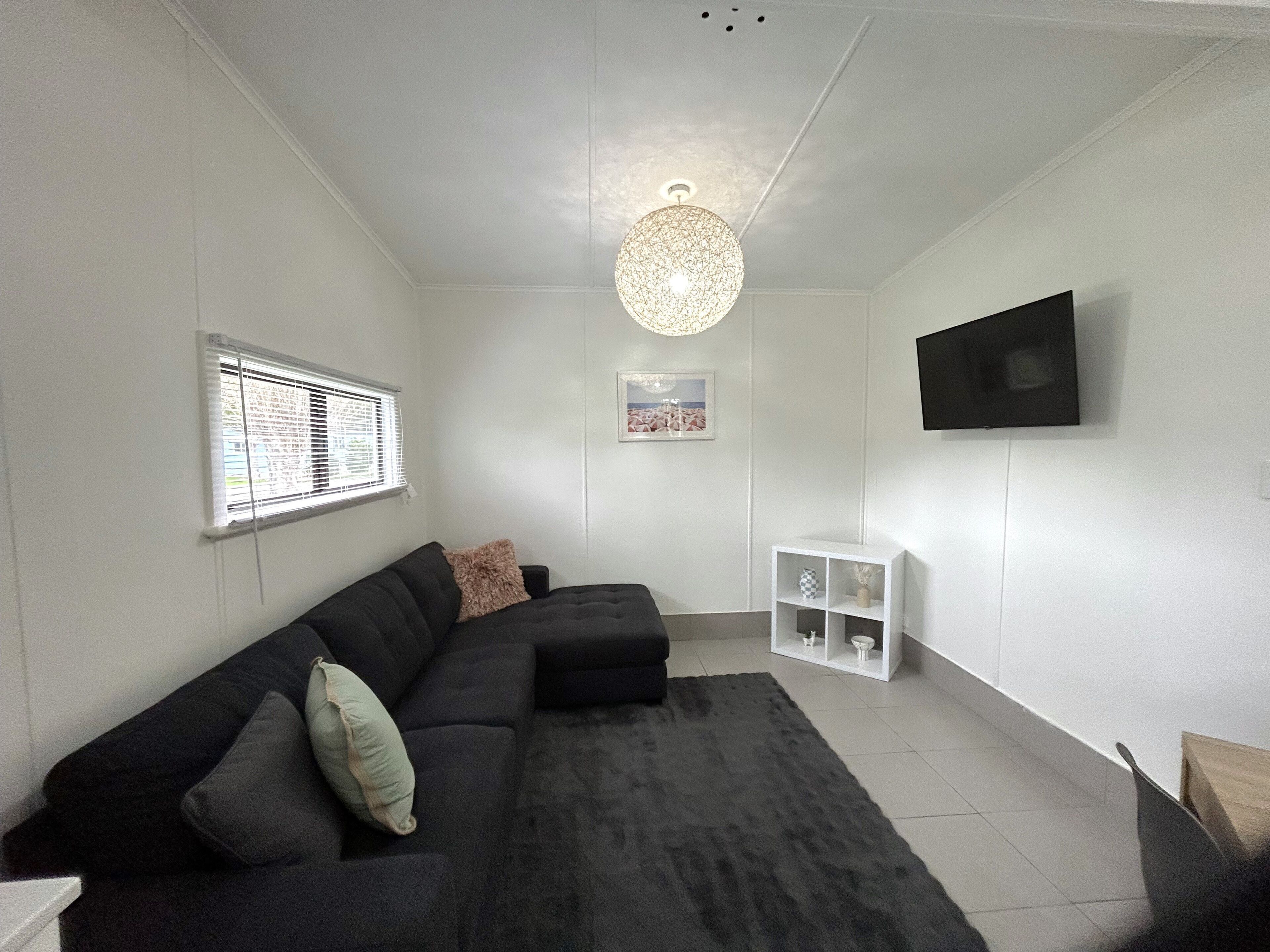 A modern living room with a dark grey sectional sofa, a grey rug, a white shelving unit, and a ceiling light fixture.