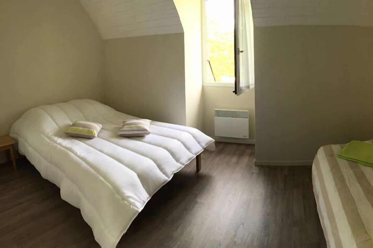 A bedroom with a bed on the left and a sofa on the right, a window with curtains, and a radiator.