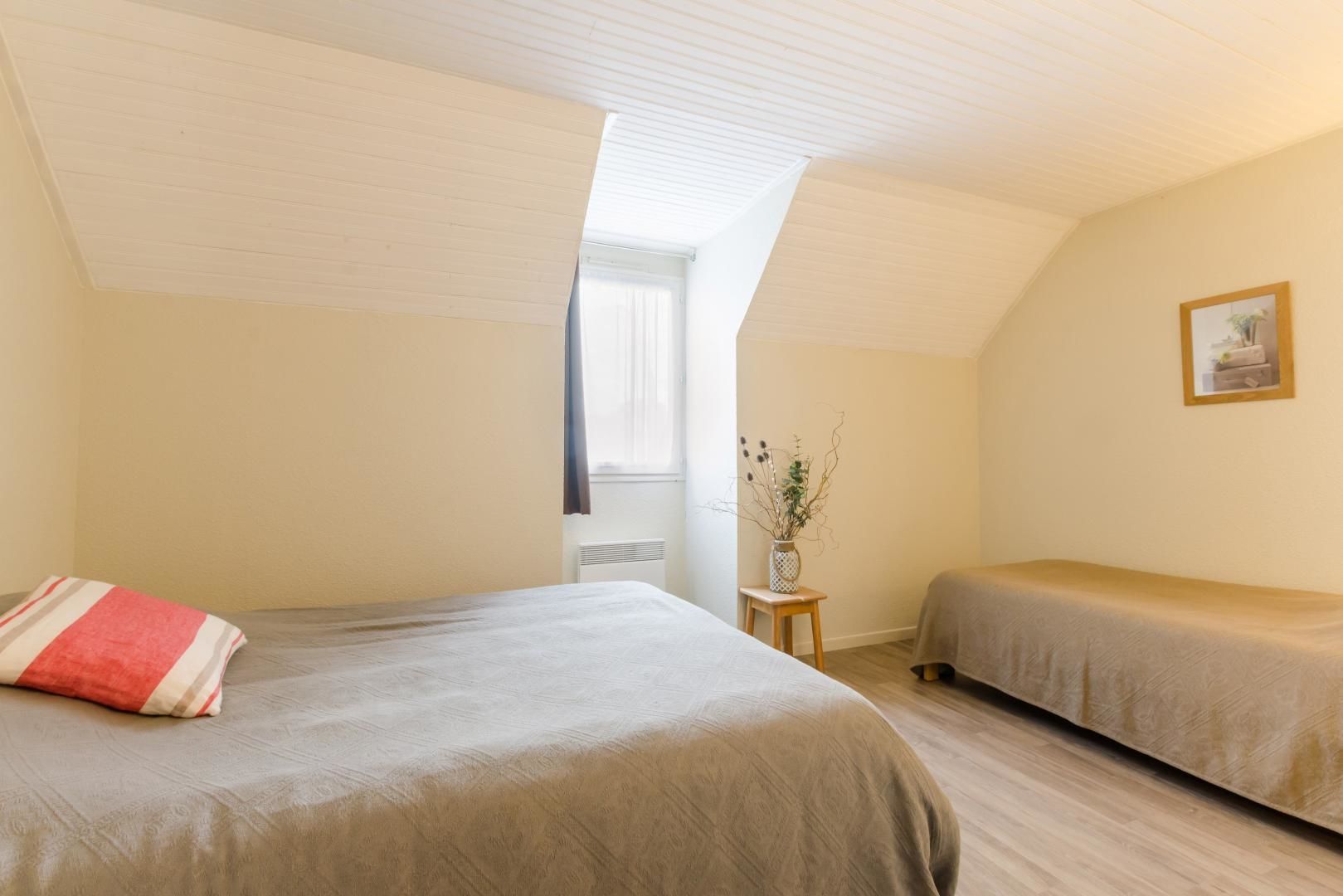 A bedroom with two beds, a small table with a vase, and a window with curtains.