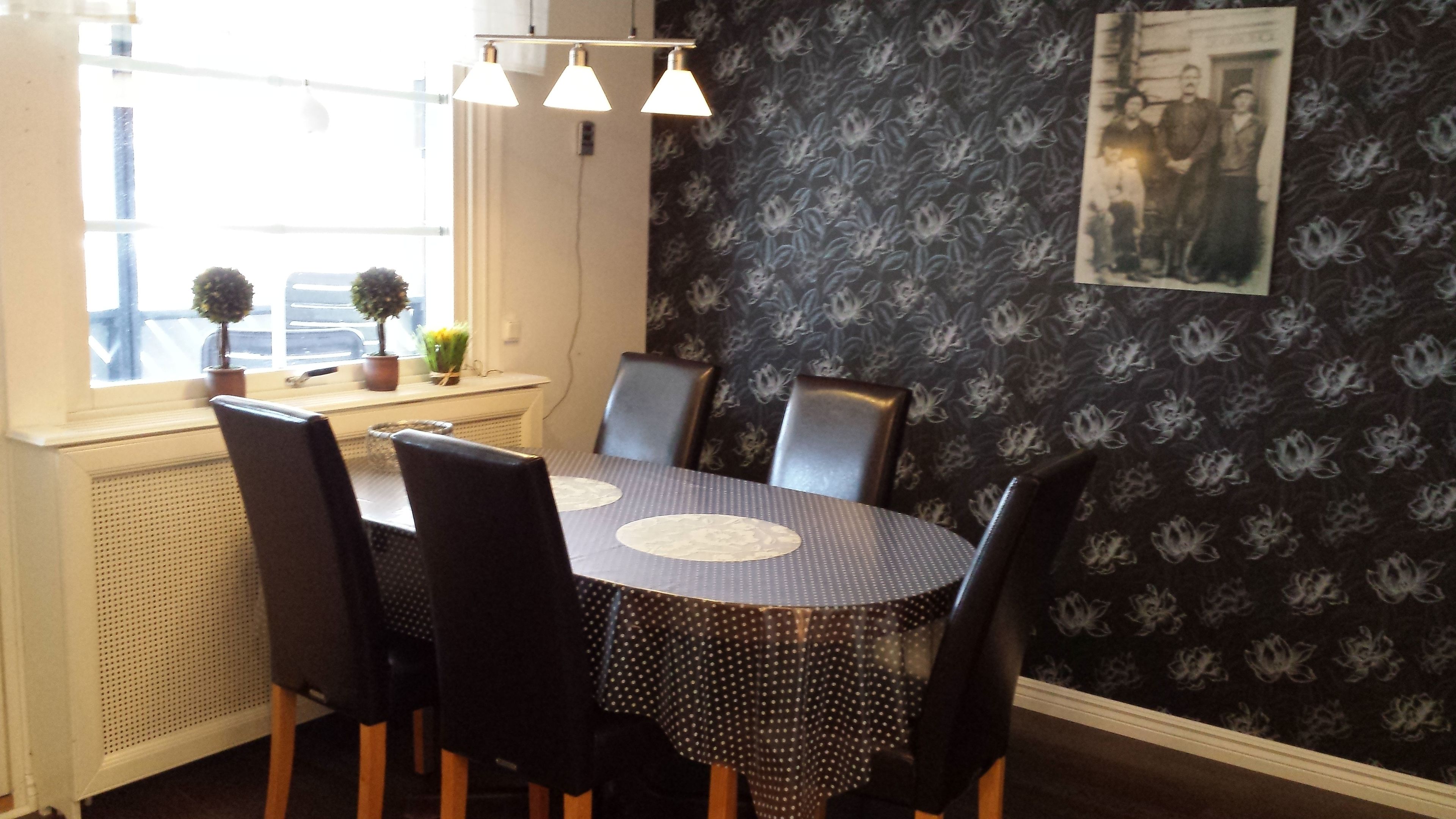 Un coin repas avec une table ronde, des chaises noires, un papier peint à motifs et une fenêtre ornée de plantes.