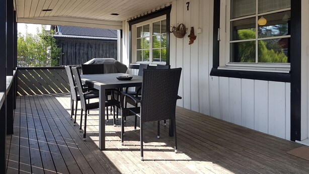 Un patio techado al aire libre con mesa y sillas, una parrilla y una ventana con vistas a la vegetación.