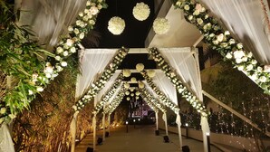 An indoor venue with a central aisle flanked by white drapes and floral arrangements, leading to a stage area. - The Emerald Club (Lodhika)