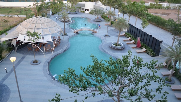 A large outdoor swimming pool with a bridge, surrounded by palm trees and lounge chairs. - The Emerald Club (Lodhika)