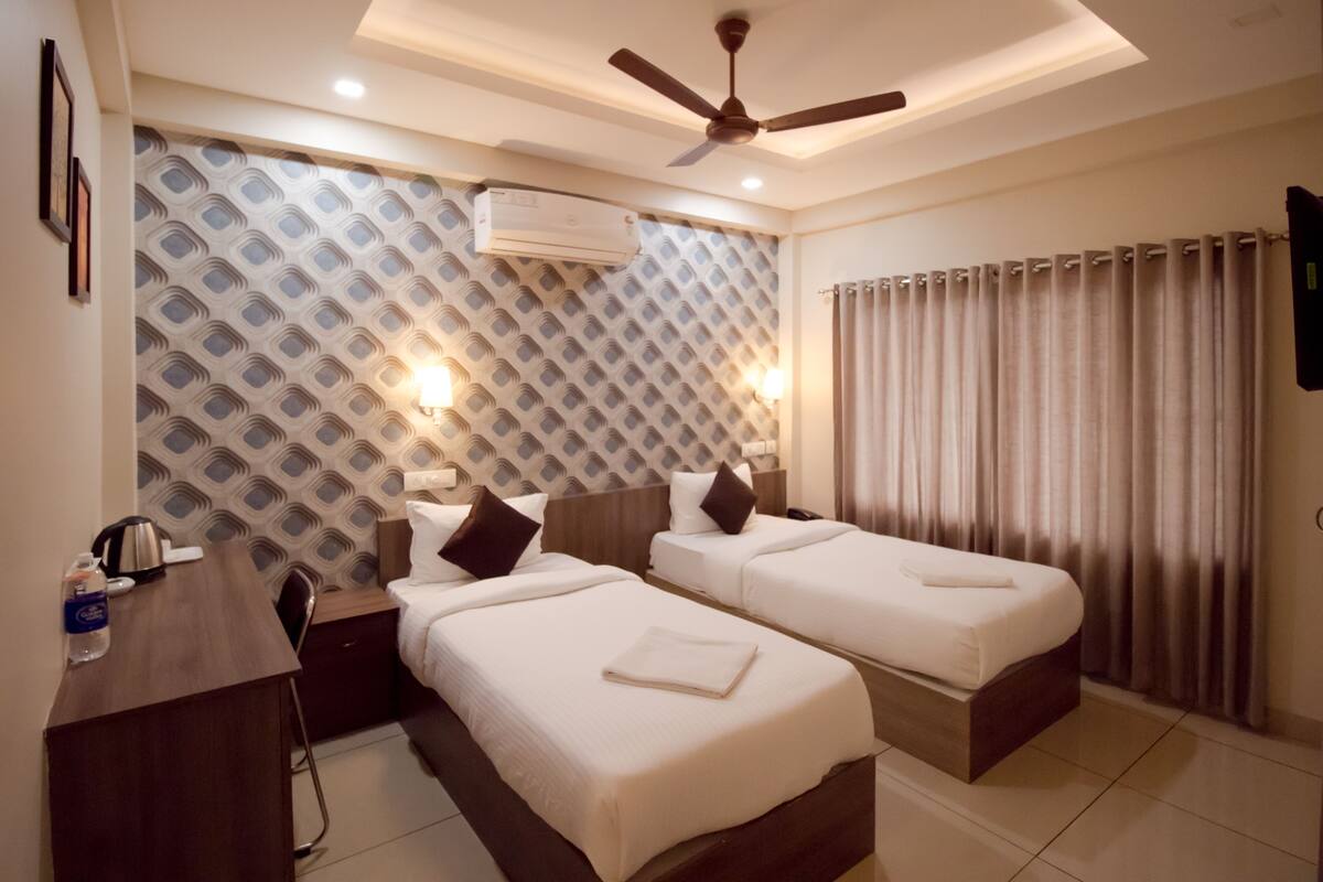 Ein Hotelzimmer mit zwei Betten, einem Schreibtisch, einer Klimaanlage, einem Deckenventilator und einem Vorhang.
