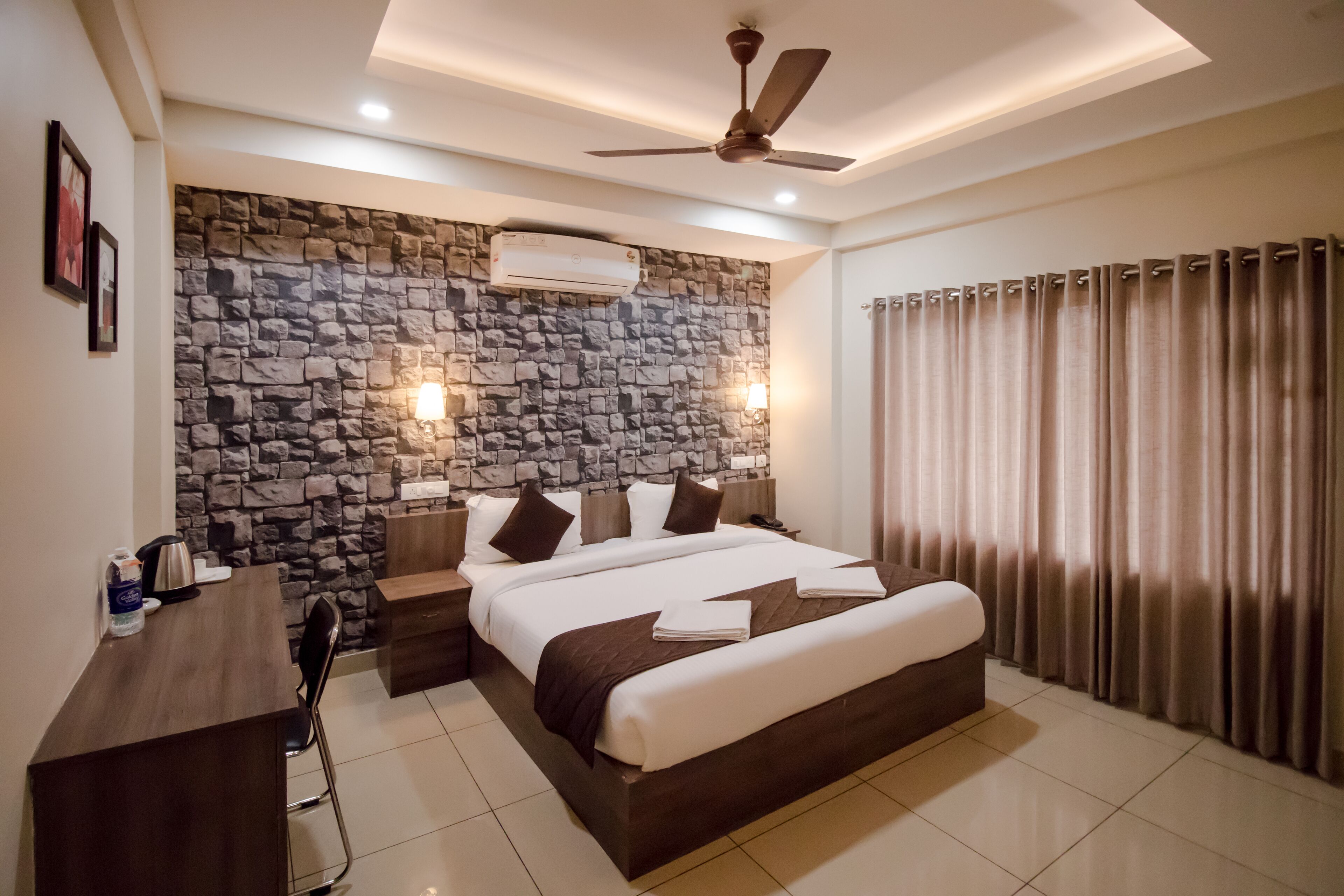 A modern hotel room with a stone wall, a large bed, a desk, and a ceiling fan.