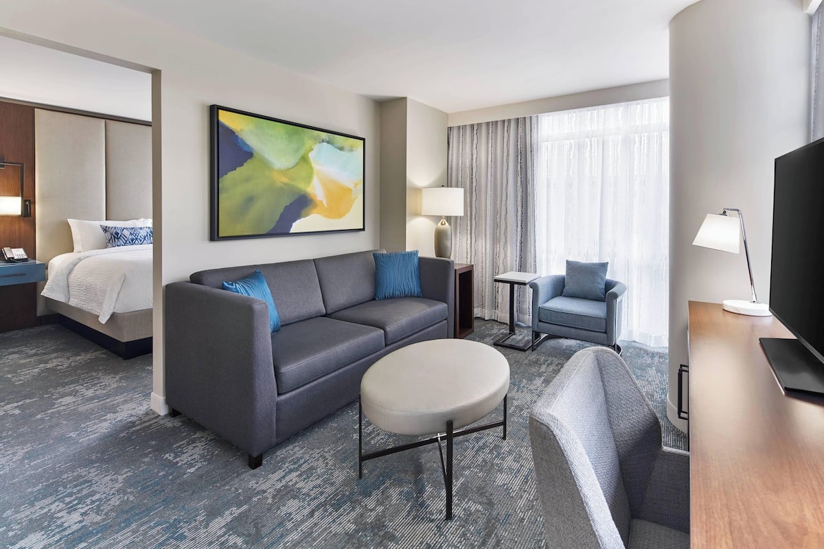 A modern hotel room with a gray sofa, a round coffee table, a TV, and a bed with a white headboard.