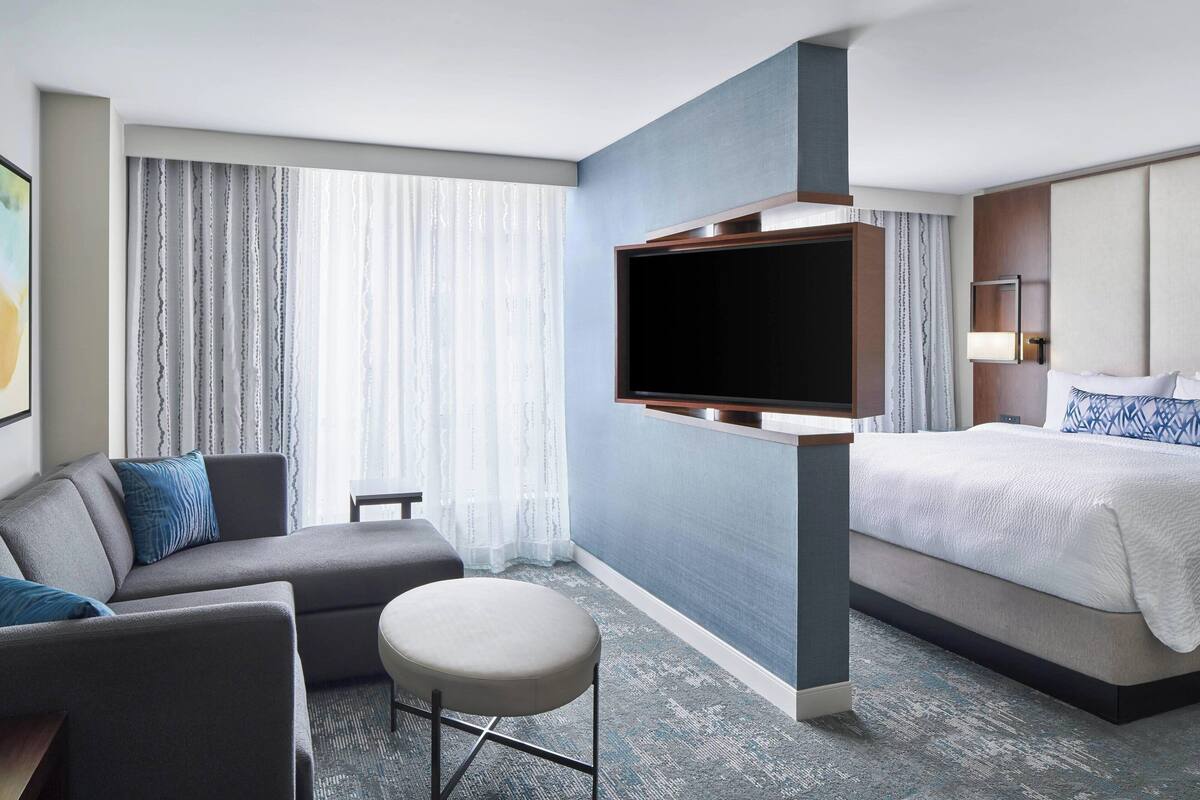 A modern hotel room with a flat-screen TV mounted on a wall, a sofa, a small table, and a bed with a headboard.