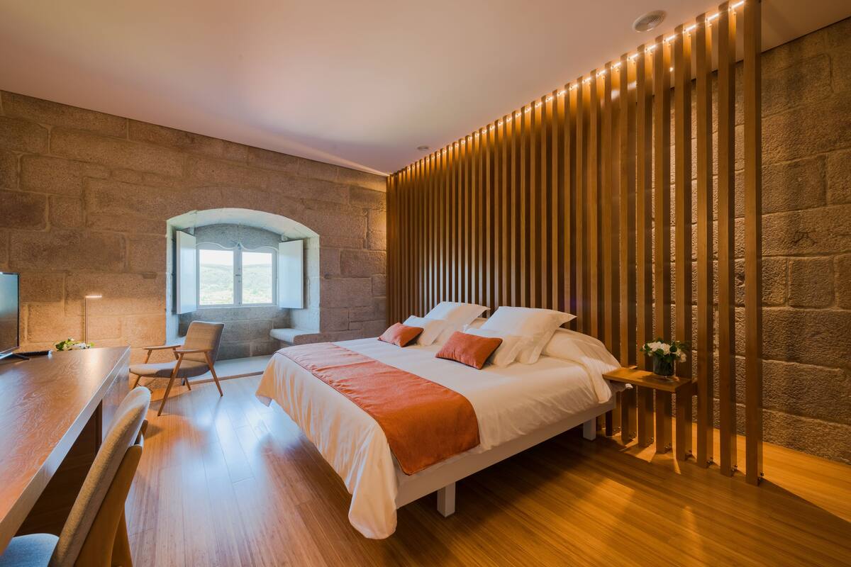 A modern bedroom with a large bed, a desk, and a chair. There is a stone wall and a wooden panel on one side.