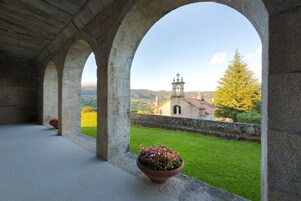 Un pasillo de piedra con arcos, una maceta y vistas de una iglesia y el paisaje.