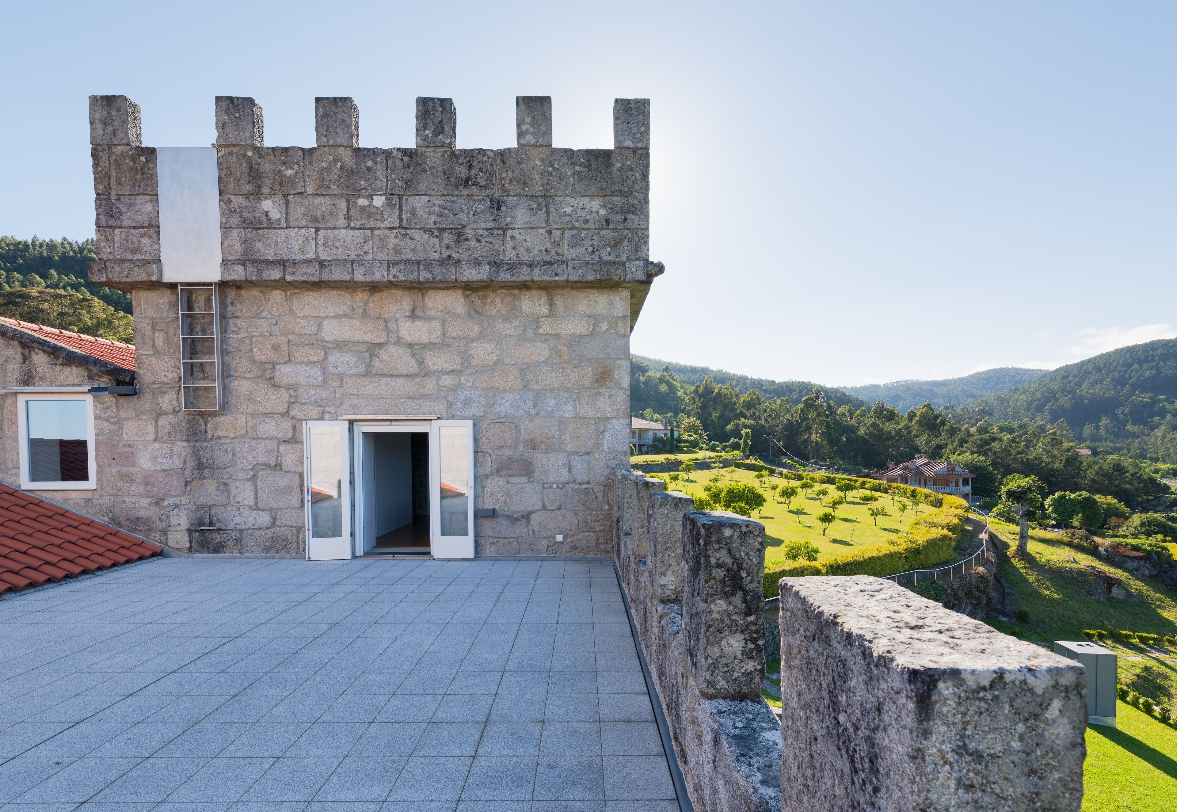Un edificio de piedra con estructura de castillo, una terraza con tejas y vistas a un paisaje verde con colinas.