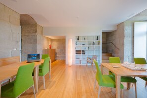 Un espace de salle à manger moderne avec des tables en bois et des chaises vertes, une bibliothèque agrémentée d’objets décoratifs et un mur en pierre.