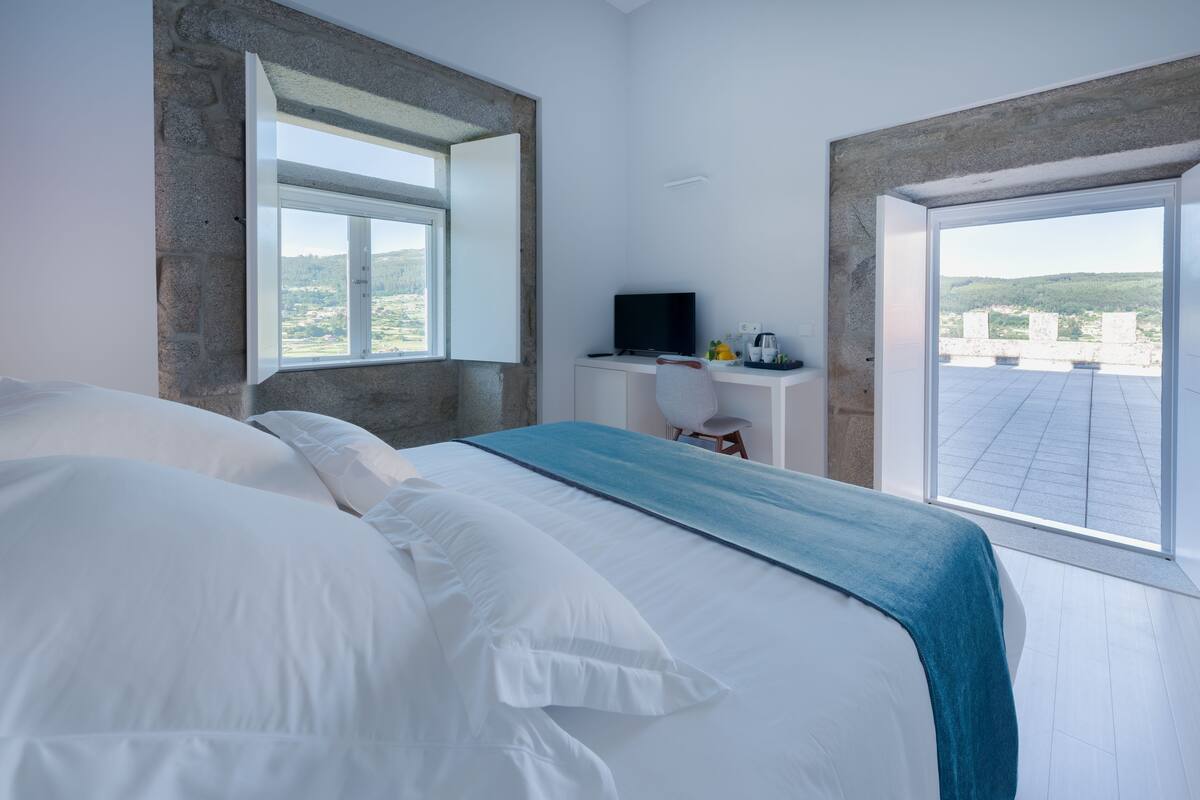 A bedroom with a large bed, a desk with a TV, and a view of the countryside.
