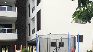 Un parque infantil con un trampolín y estructuras para escalar, rodeado de zonas verdes cuidadas y un edificio con balcones.