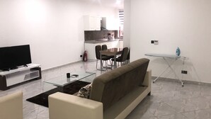 Una sala moderna con sofá, una mesa de centro y televisor. Se observa un comedor con sillas y una mesa, y una cocina al fondo.