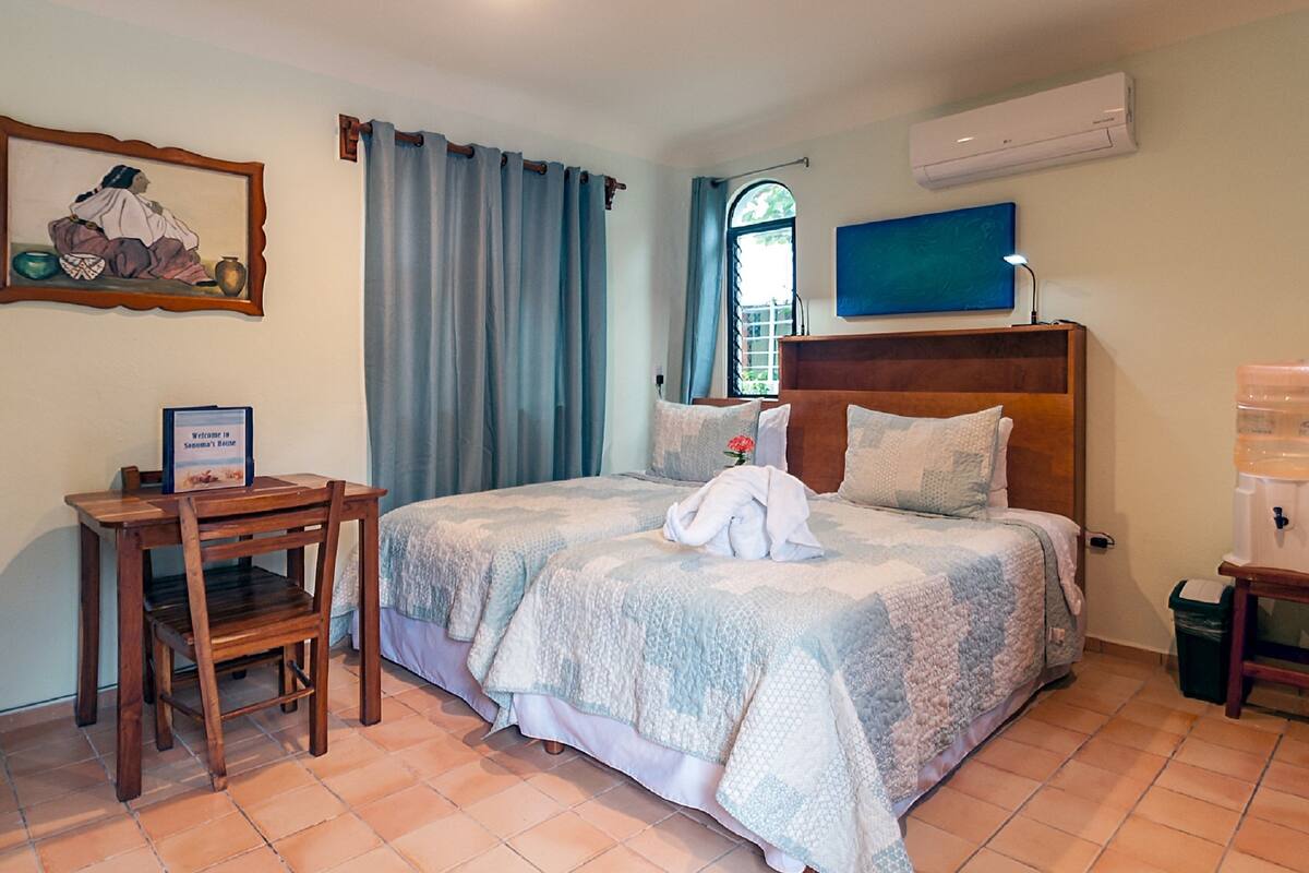 A bedroom with a bed, a wooden desk, a framed picture, and a window with curtains.