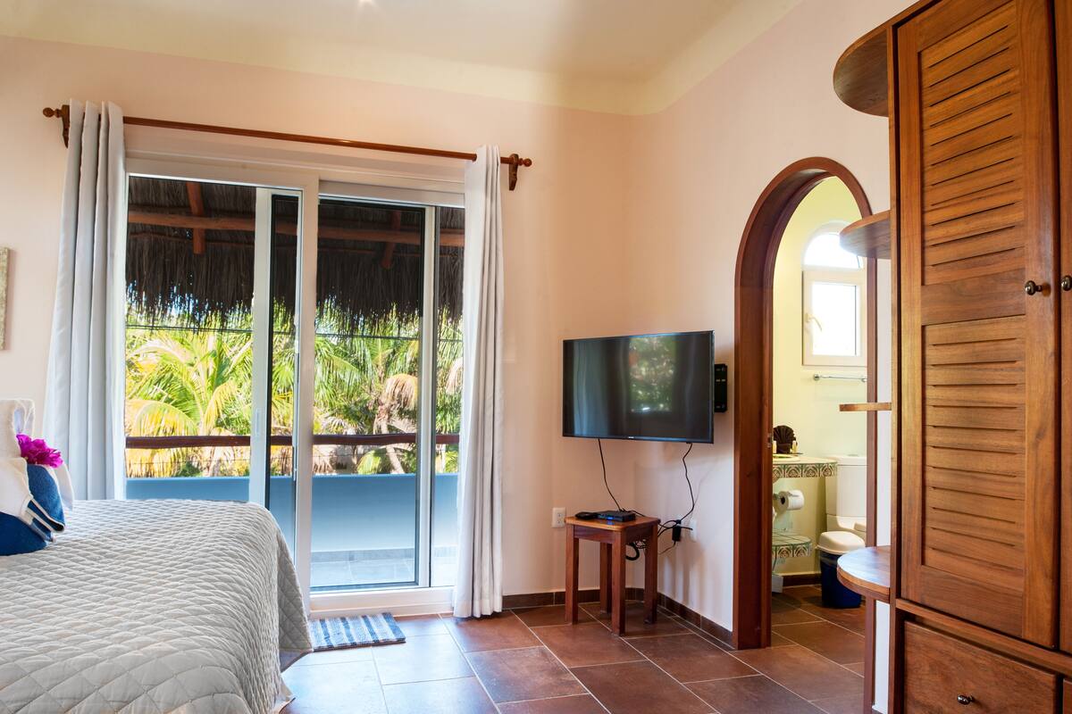 A bedroom with a bed, a TV, a sliding glass door to a balcony, and a wooden wardrobe.