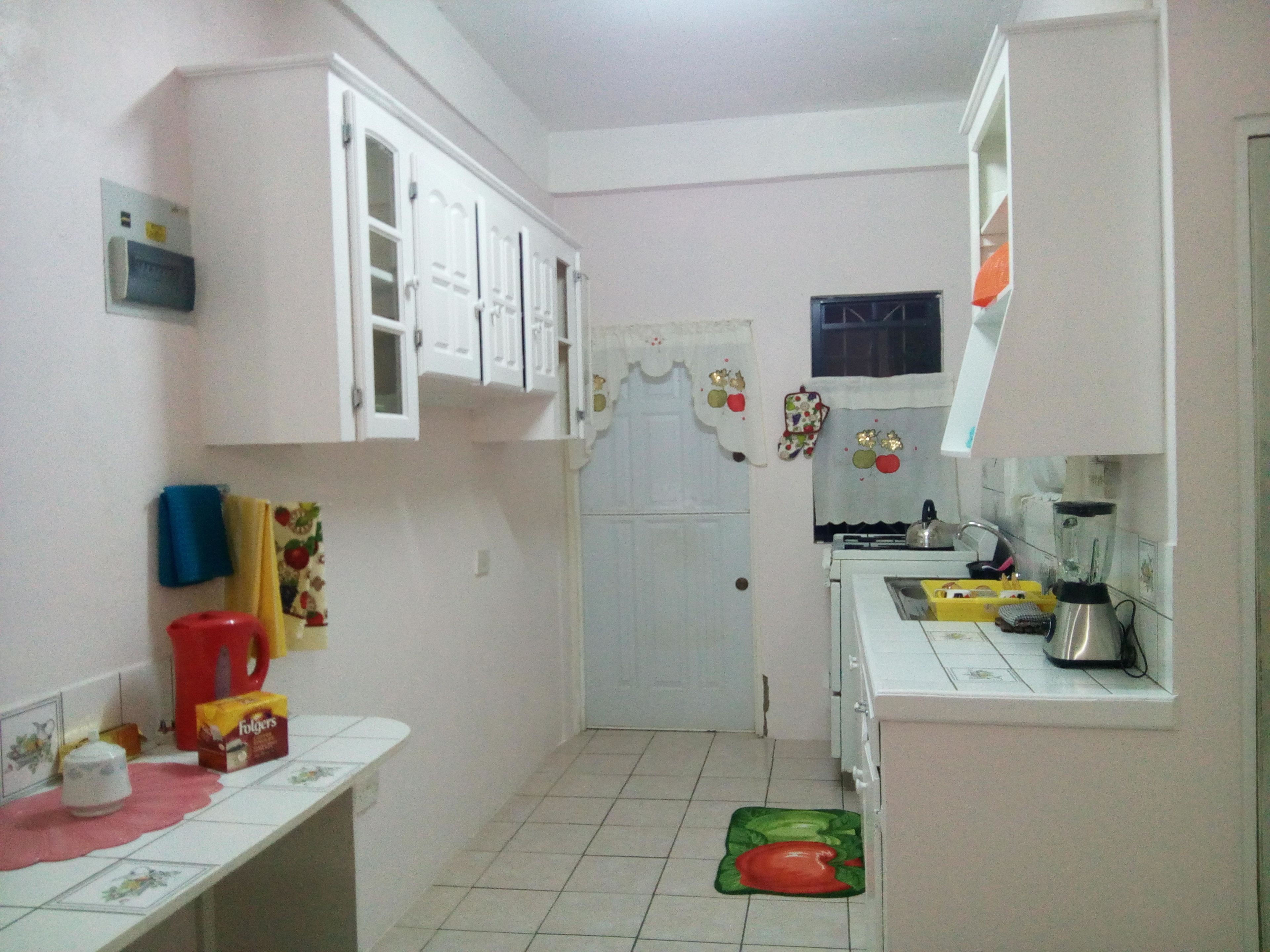 A small kitchen with white cabinets, a refrigerator, a sink, and a stove. There is a window with a curtain and a door with a decorative wreath.